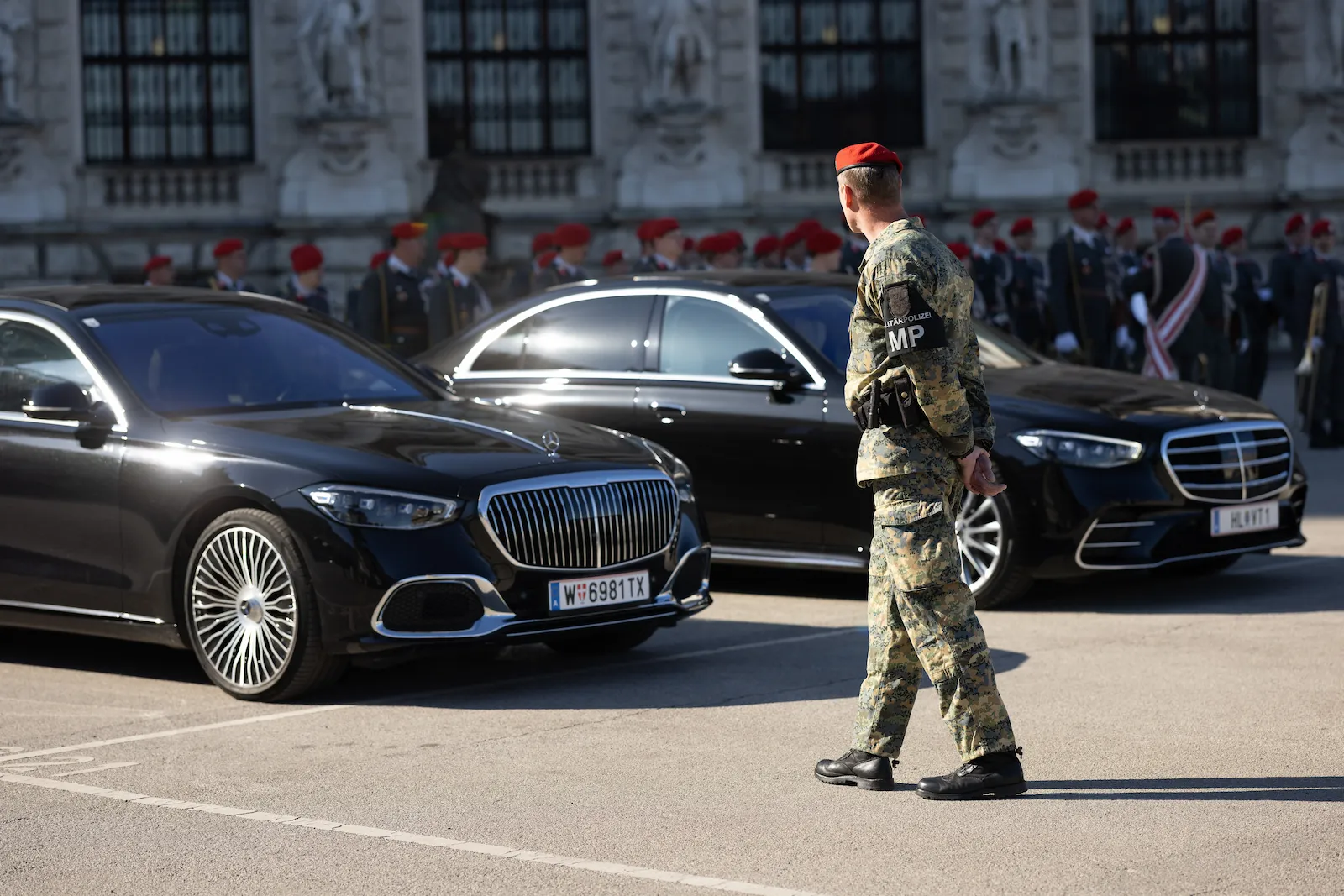 Der Diplomaten Fahrdienst zeigt schwarze Luxuslimousinen mit einem bewaffneten Offizier vor einem historischen Gebäude.