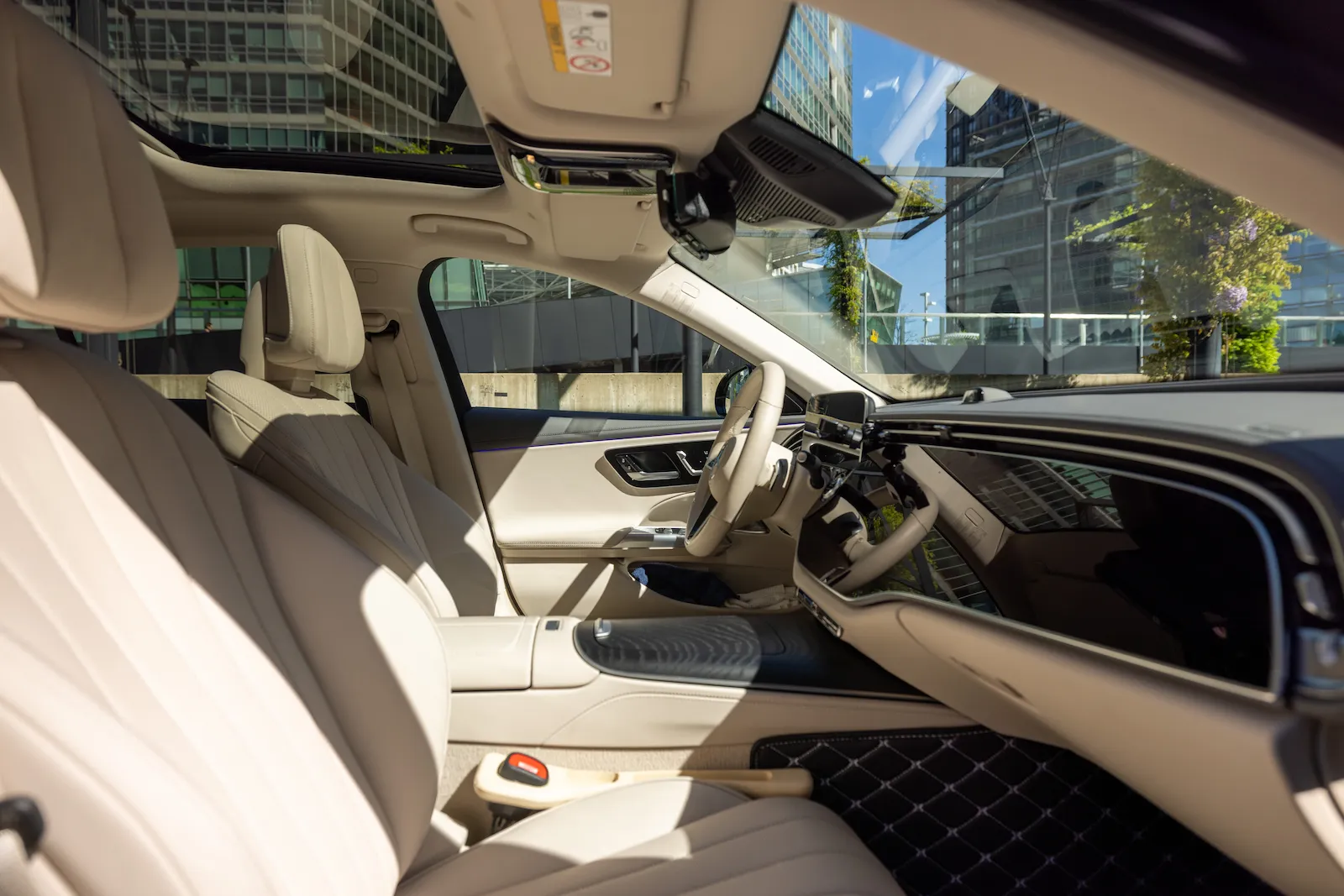 Luxuriöse Mercedes S-Klasse Innenansicht mit beigem Lederinterieur, Lenkrad, Armaturenbrett und Panoramadach vor städtischem Hintergrund.