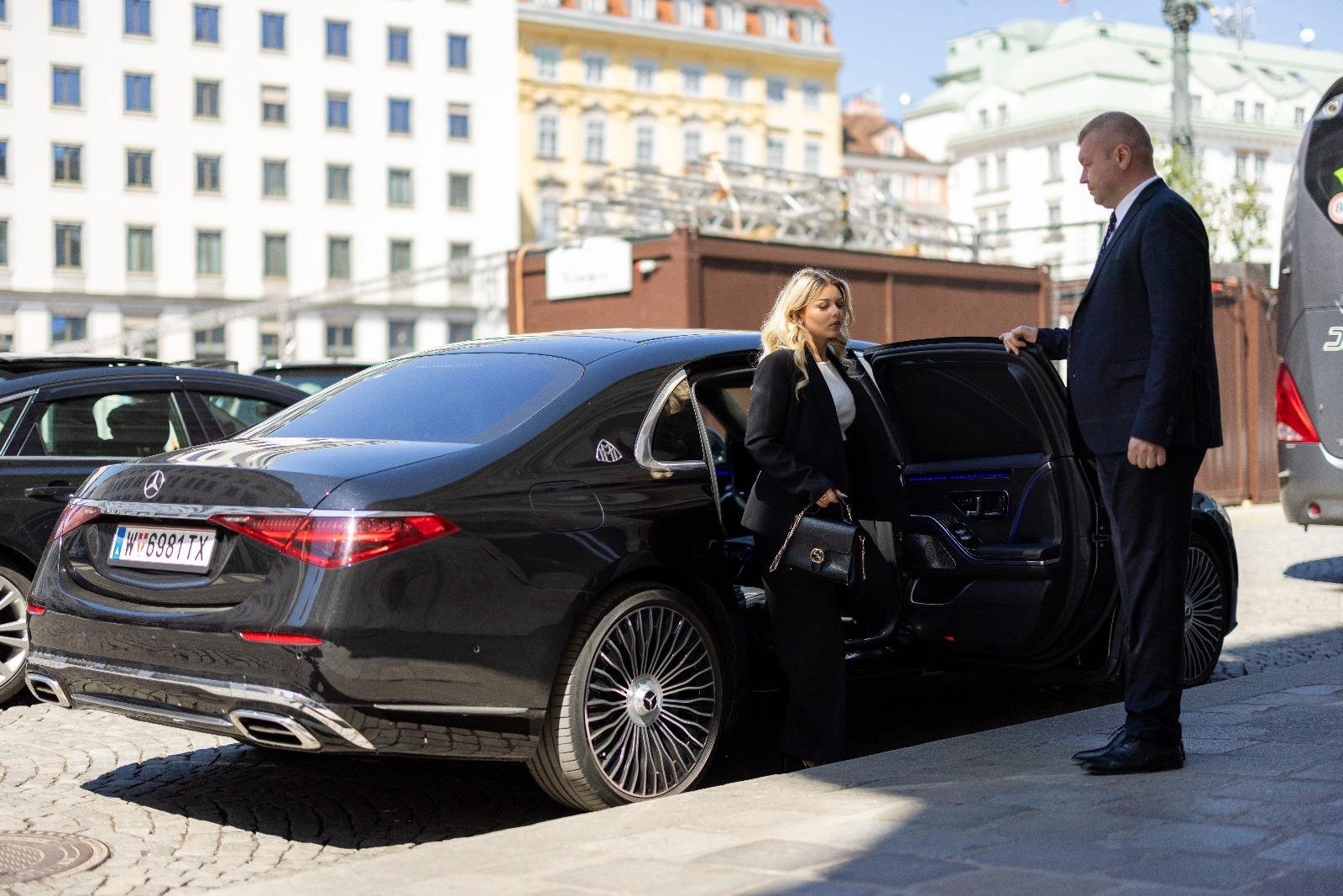 Ein Luxus Chauffeurservice mit einem schwarzen Mercedes, bei dem ein Chauffeur einer eleganten Dame die Tür öffnet vor Gebäude.